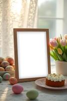 A blank wooden frame sits on a table adorned with Easter eggs, a festive Easter cake, and a vase of tulips, creating a warm and inviting springtime scene photo