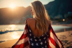 Young blonde woman covered her shoulders with the American flag of the USA and look at the bay of the ocean, view from the back photo