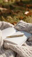 cozy scene featuring blank notebook and gold pen resting on knitted blanket, surrounded by autumn leaves, evokes sense of warmth and creativity photo