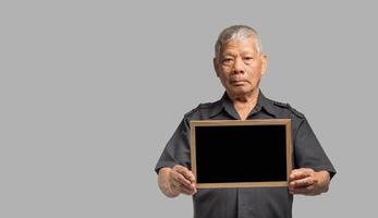 Portrait of an elderly Asian man holding a mini blackboard and looking at the camera with a smile while standing on a gray background. photo