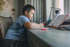 asian boy write in notebook, homework beside open textbook on desk, study tool, education, homeschooling, parenting resources photo