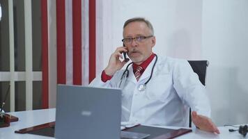 Senior physician consultation using mobile phone. Professional doctor in medical coat and stethoscope sitting in his cabinet and giving a conversation through the phone with a patient. video