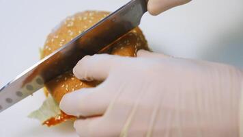 Hands in gloves of a cook making burger. Chef cutting ready hamburger into halves with a sharp knife on table. Close-up. video
