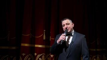 Speaker on stage. Male host in elegant costume speaking into a microphone looking on camera on the background of red curtains in the theater. Slow motion. video