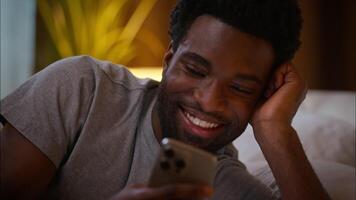Close up laughing happy African American man male guy user holding mobile phone lying in bed scrolling social media laugh cellphone online addicted gadget chatting bedtime at home bedroom late night video