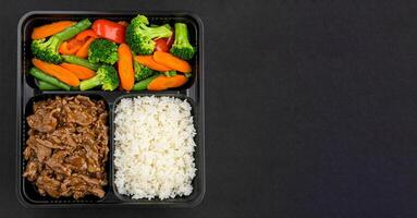 Black bento Box container holds balanced meal of beef stir-fry with vegetables, and white rice in separate compartments. Container is placed on black table background, top view, copy space for text. photo