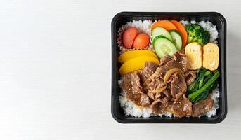 Black bento Box container holds balanced meal of beef stir-fry with vegetables, and white rice in separate compartments. Container is placed on light table background, top view, copy space for text photo