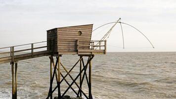 Traditional French fishing hut on stilts with carrelet net over ocean. Wooden structure stands on rocky shore, with waves and cloudy sky. Ideal for projects to travel, close-up photo