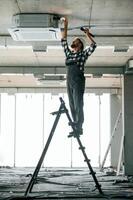 Standing on, the ladder. Repairman in uniform is in the unfinished building photo