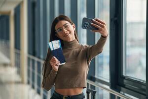 Making selfie ,with phone, smiling. Young woman traveler in the airport entrance hall photo