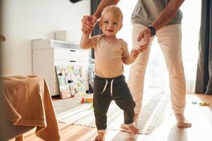 Boy is ,,walking by the help of dad. Man is playing with his little son in the domestic room photo