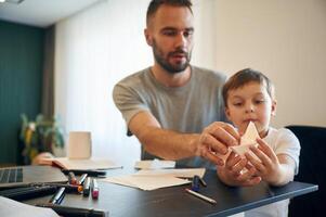 Playing with,, origami boat. Father and son are at home by the table photo