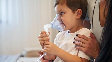 Boy is ,making, inhalation with nebulizer to reduce coughing. Mother with her little son is at home in domestic room photo