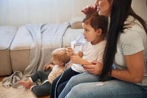 Boy is making inhalation with nebulizer, to reduce coughing. Mother with her little son is at home in domestic room photo