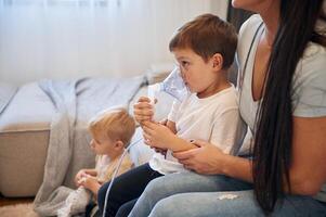 Boy, is making, inhalation with nebulizer to reduce coughing. Mother with her little son is at home in domestic room photo