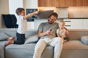 Pillow ,push by the kid. Father with two baby boys is sitting at sofa photo