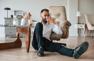 sentado en el piso, triste. trastornado joven hombre bebedor alcohólico es adentro foto