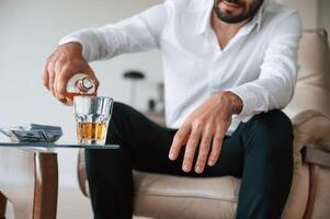 Pouring, the whiskey into the glass. Upset young man drinker alcoholic is indoors photo