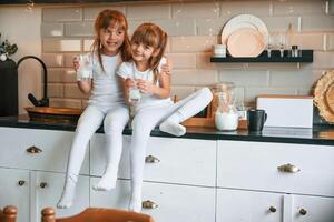Embracing each other. Happy two little girls are drinking milk while sitting on the table on the kitchen photo