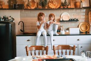 Having fun. Happy, two, little girls are drinking milk while sitting on the table on the kitchen photo