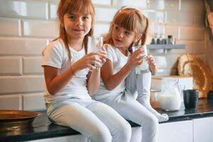 Having fun. ..Happy two little girls are drinking milk while sitting on the table on the kitchen photo