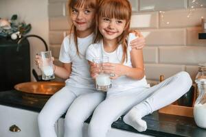Embracing each other. Happy, two little girls are drinking milk while sitting on the table on the kitchen photo