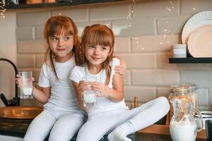 Embracing each ,other. Happy two little girls are drinking milk while sitting on the table on the kitchen photo