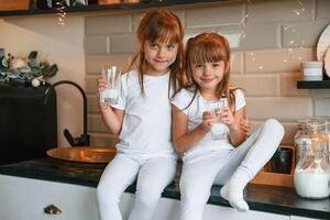 Embracing, each other. Happy two little girls are drinking milk while sitting on the table on the kitchen photo
