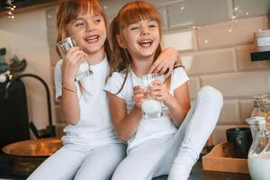 Happy, two little girls are drinking milk while sitting on the table on the kitchen photo