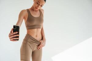 Taking a, photo with phone. Woman in fitness clothes is standing against white background