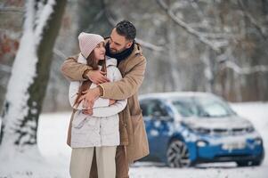 Standing ,and embracing.. Happy couple having a walk in winter forest. Blue car is parked photo