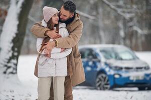 Standing ,and ,embracing. Happy, couple having a walk in winter forest. Blue car is parked photo