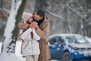 Standing and embracing... Happy couple having a walk in winter forest. Blue car is parked photo