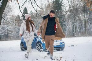 Front view, having fun. Happy couple having, a walk in winter forest. Blue car is parked photo
