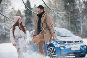 Man is ,kicking the ,snow. Happy couple having a walk in winter forest. Blue car is parked photo