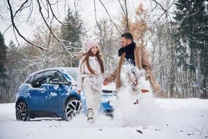Man is, kicking, the snow. Happy couple having a walk in winter forest. Blue car is parked photo