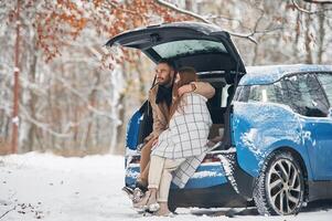 Leaning on the automobile... Happy couple having a walk in winter forest. Blue car is parked photo