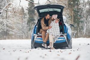 Leaning on the, automobile. Happy ,couple having a walk in winter forest. Blue car is parked photo