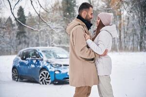 Standing ,and, embracing each other. Happy couple having a walk in winter forest. Blue car is parked photo