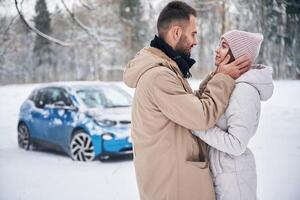 Standing, and, embracing each other. Happy couple having a walk in winter forest. Blue car is parked photo