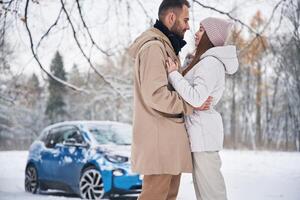 Standing and, embracing each other. Happy, couple having a walk in winter forest. Blue car is parked photo
