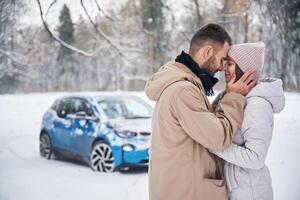 Standing,, and embracing each other. Happy couple having a walk in winter forest. Blue car is parked photo