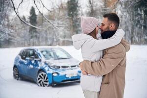 Standing, and, embracing, each other. Happy couple having a walk in winter forest. Blue car is parked photo