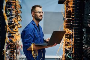 With laptop in hands,. Young, man is working with internet equipment and wires in server room photo
