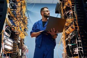 Holding ,laptop. Young, man is working with internet equipment and wires in server room photo