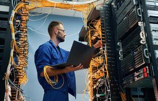 With laptop in hands. Young man, is, working with internet equipment and wires in server room photo