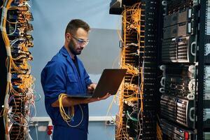 With laptop in hands,. Young man is working with internet equipment and wires in server room photo