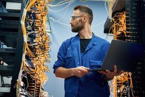 Holding laptop. Young, man, is working with internet equipment and wires in server room photo