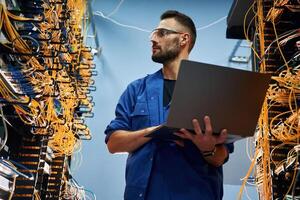 Holding, laptop. Young man is working ,with internet equipment and wires in server room photo