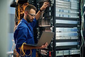 Using the laptop. Young, man, is working with internet equipment and wires in server room photo
